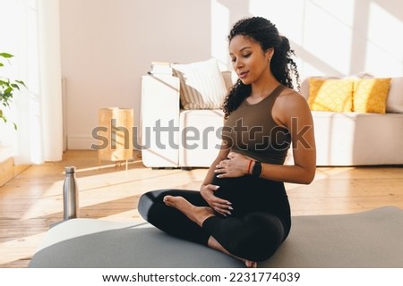 Pregnant african woman in sports clothes sitting on mat in her living-room, having rest after doing prenatal fitness training exercise, stroking her belly, feeling pushes of her baby Royalty-Free Stock Photo #2231774039