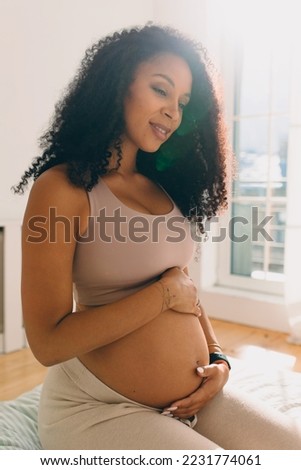 Vertical image of gentle pregnant woman with dark skin and curly hair sitting on bed in the morning, touching her belly, communicating with her child, pushing her from the inside. Happy pregnancy