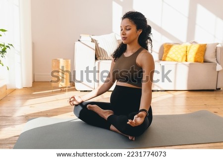 Charming pregnant african american woman in leggings and crop top sitting on floor in living-room in lotus posture with closed eyes, doing mudra sign with fingers, reaching zen, balance and harmony Royalty-Free Stock Photo #2231774073