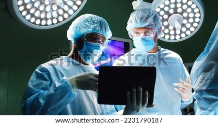 Close up portrait of mixed-races doctors male surgeons speaking and typing on tablet while standing in operating room performing operation on patient under the bright lamps in hospital during surgery Royalty-Free Stock Photo #2231790187