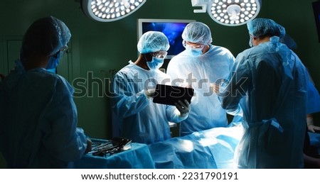 Multi-ethnic doctors professional surgeons talking and browsing on tablet standing in operating room in hospital performing operation on patient under bright lamps during surgery. Emergency concept Royalty-Free Stock Photo #2231790191