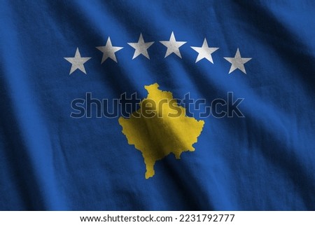 Kosovo flag with big folds waving close up under the studio light indoors. The official symbols and colors in fabric banner