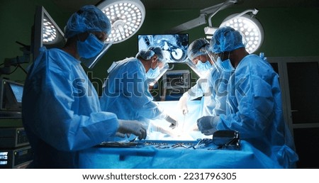 Team of professional mixed-races doctors and assistants in medical uniforms performs heart transplant operation under lamp using medical tools in operating room looking at monitor. Surgery concept Royalty-Free Stock Photo #2231796305