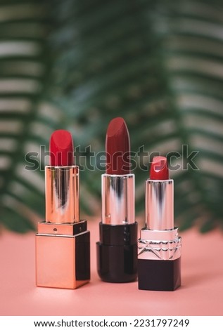 Three pieces of red lipstick stands on a pink table with palm branches in the background, close-up side view with depth of field. The concept of cosmetics, beauty salon.