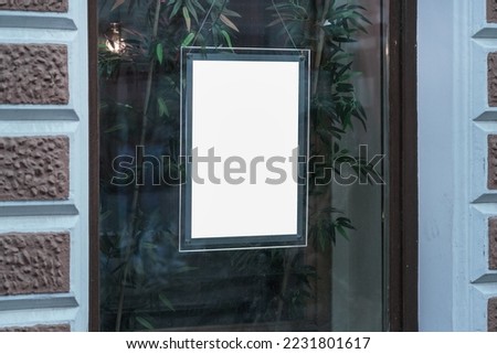 Empty white poster in glass frame fixed on window of street cafe or shop. Blank signboard for advertising information.