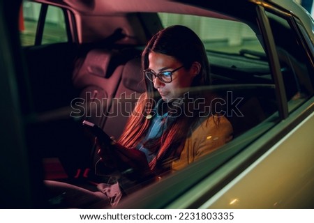 Businesswoman traveling with car on a business trip during night while sitting in a backseat and using a smartphone. Female using mobile phone to send email or messages. View trough car window. Royalty-Free Stock Photo #2231803335