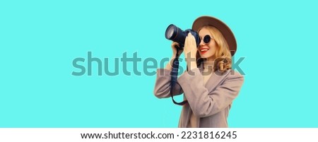 Portrait of stylish happy smiling woman photographer with digital camera taking picture wearing round hat, brown coat on blue background, blank copy space for advertising text