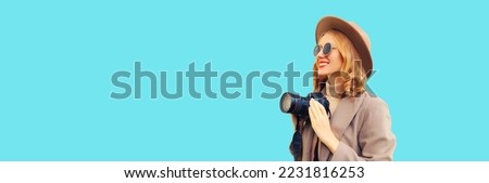 Portrait of stylish happy smiling woman photographer with digital camera taking picture wearing round hat, brown coat on blue background, blank copy space for advertising text