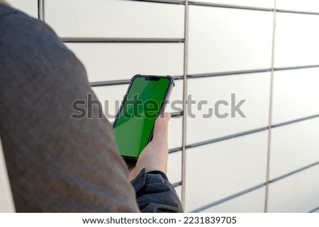 Female hand holding mobile phone with green screen chroma key next to Parcel locker Collecting parcel from shopping locker. Woman Skans QR Code on Mobile phone Self-service Locker Cell with Bar code