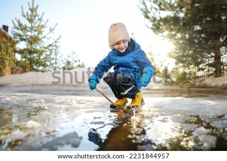 Preschooler boy is playing with a stick in brook on sunny day. Child having fun and enjoy a big puddle. All kids love play with water. Happy childhood. Outdoor activity for baby in early sping time