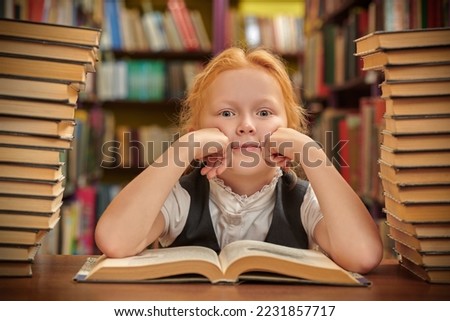 A dreamy schoolgirl in uniform sits in a school library among piles of books smiling. Modern children and education.