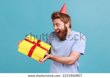 Portrait of extremely happy bearded man being very glad to get present from friend or girlfriend, holding gift box, expressing excitement. Indoor studio shot isolated on blue background.