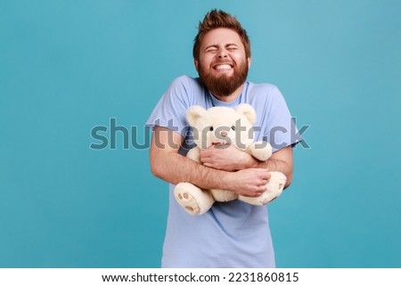 Portrait of satisfied handsome bearded man standing with closed eyes and toothy smile, embracing white soft teddy bear, expressing positive emotions. Indoor studio shot isolated on blue background. Royalty-Free Stock Photo #2231860815