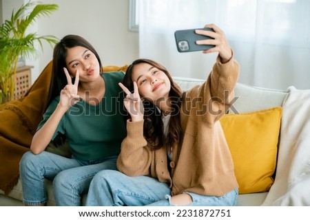 Two asian young women happy smiling and taking selfie on couch in living room at home