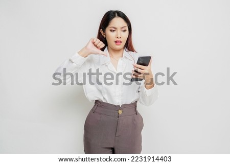 A disappointed Asian woman wearing white shirt gives thumbs down hand gesture of disapproval, isolated by white background