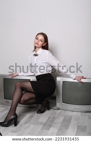 Fashion concept image of stylish girl in white shirt and black skirt posing in minimalist room with old vintage soviet times tv set.