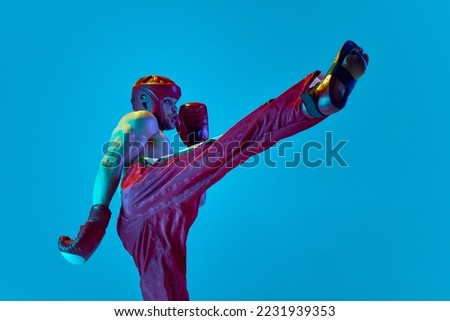 Energetic workout of professional athlete. Kickboxer in helmet and gloves training isolated over blue background in neon light. Muay thai. Sport, competition, energy, combat sports