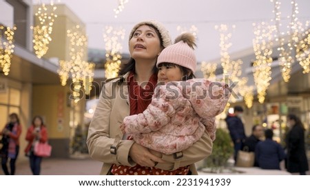 smiling asian mother and baby turning and looking around at beautiful outdoor Christmas lights above head at a mall while shopping for the winter holiday