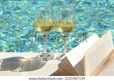 Glasses of tasty wine and open book on swimming pool edge