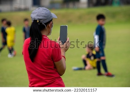 Mom standing and taking pictures  her son playing football in a school tournament on a sideline with a sunny day. Sport, outdoor active, lifestyle, happy family and soccer mom and soccer dad concept.