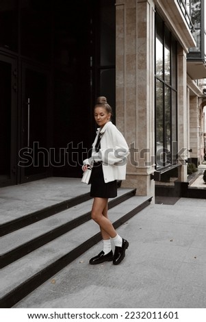 beautiful young woman dressed in black skirt and top, white furry jacket, patent leather loafers, socks, white bag, bun hairstyle, stylish fashion outfit, full length lifestyle model Royalty-Free Stock Photo #2232011601