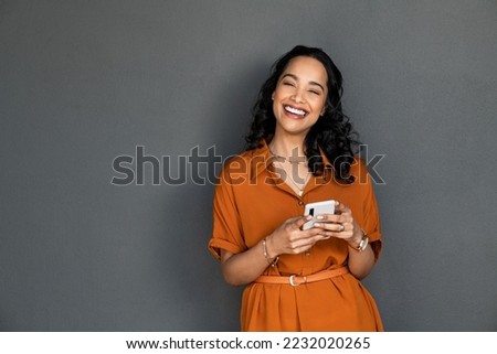 Portrait of beautiful mixed race woman standing against grey wall using smartphone. Cheerful latin young woman holding mobile phone while looking at camera. Happy carefree woman messaging.
