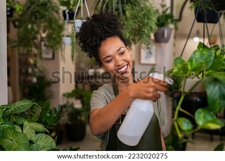 Beautiful black florist taking care of plants while spraying it with water. African american owner working and spraying water plants in store. Happy and smiling florist watering plants in shop. Royalty-Free Stock Photo #2232020275