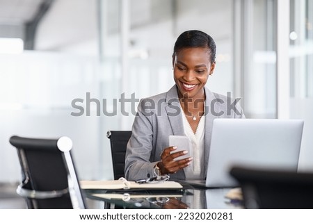 African american businesswoman using smartphone while working on laptop in office. Smiling mature black business woman checking phone while working. Successful woman entrepreneur browsing on phone.
