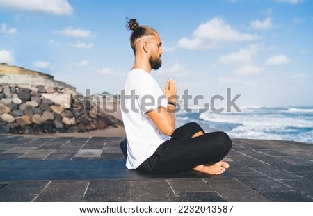 Low angle side view of young male seeking holistic spirituality sitting in Namaste Mudra on stone structure in front of sea while meditating with closed eyes for appeasement and mindfulness Royalty-Free Stock Photo #2232043587