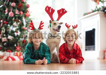 Christmas at home. Kids and dog in reindeer antlers under Xmas tree. Little child hug puppy in deer hat and open Christmas presents. Children play with pet. Winter holiday celebration. 