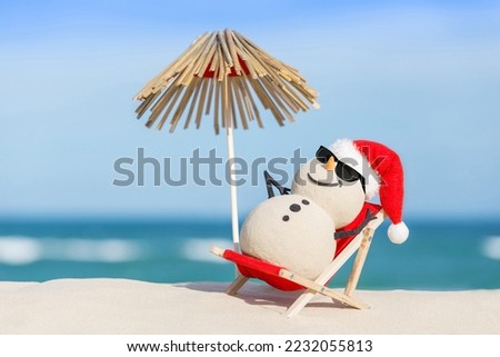 Sandy Christmas Snowman is enjoying the sun on a lounge next to a thatched umbrella, wearing sunnies