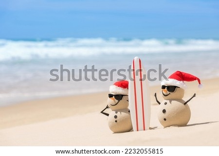 Two Sandy Christmas Snowmen are watching the waves, standing on beautiful beach with a surf board, wearing sunnies