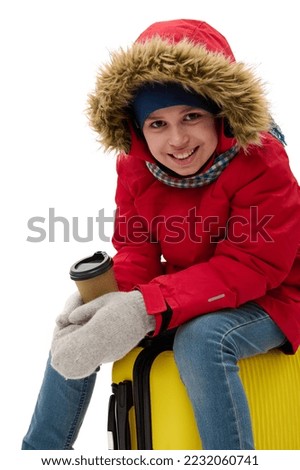 Happy traveler child boy in red down jacket, blue jeans, wool mittens smiling a toothy smile to camera, sitting on suitcase with a disposable cup of hot drink. Winter holidays. Travel. Journey concept
