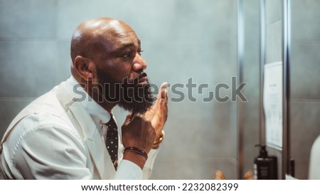 A profile view of a handsome bald black man grooming, brushing, and moisturizing the beard hair in front of the mirror in a bathroom of a luxury hotel; an African-American man taking care of his beard Royalty-Free Stock Photo #2232082399
