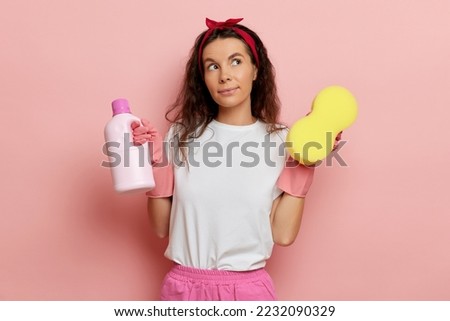 Happy and pleased housewife, holds cleaning liquid and sponge and look a side, isolated over pink wall