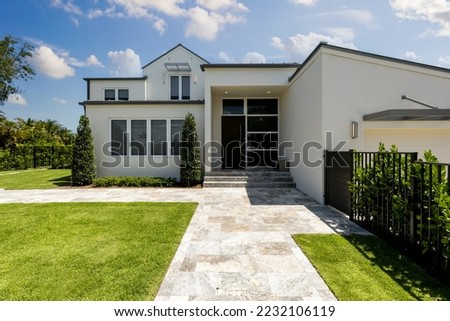 Front view of a modern stylish white house with walkway to front door, garage, short grass, trees, palms, privet fringe, sidewalk, blue sky Royalty-Free Stock Photo #2232106119