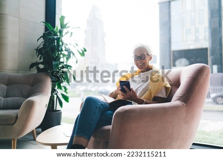Smiling aged female employee in casual outfit and spectacles reading messages on mobile phone while sitting in comfortable armchair in rest room during break
