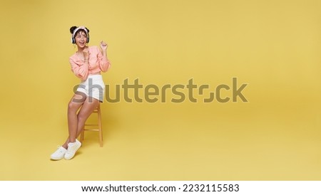 Happy young asian teen woman smiling and sitting on chair her looking and pointing to isolated on yellow background.