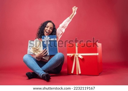Portrait of smiling latin woman with curly hair wearing sweater with gift boxes isolated on red background.