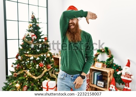 Redhead man with long beard wearing christmas hat by christmas tree covering eyes with arm smiling cheerful and funny. blind concept. 