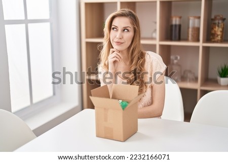 Beautiful blonde woman with cardboard box thinking concentrated about doubt with finger on chin and looking up wondering 