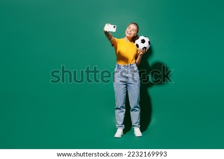 Full body young woman fan wear yellow t-shirt cheer up support football sport team hold in hand soccer ball watch tv live stream do selfie shot on mobile cell phone isolated on dark green background