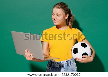 Young woman fan wearing basic yellow t-shirt cheer up support football sport team hold in hand soccer ball watch tv live stream use work on laptop pc computer isolated on dark green background studio