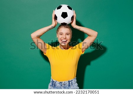 Young smiling fun woman fan wear basic yellow t-shirt cheer up support football sport team hold in hand soccer ball above head look camera watch tv live stream isolated on dark green background studio Royalty-Free Stock Photo #2232170051
