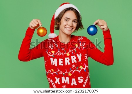 Merry young woman wear knitted xmas sweater Santa hat posing hold in hands two toys for Christmas tree isolated on plain pastel light green background. Happy New Year 2023 celebration holiday concept
