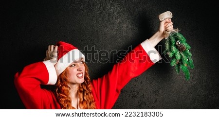 Angry crazy red-haired woman in a suit and a red Santa Claus hat holds a inverted Christmas tree in her hands on a black dark background.