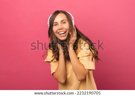 Laughing woman is having fun and listening to the music through headphones. Studio shot over pink background.