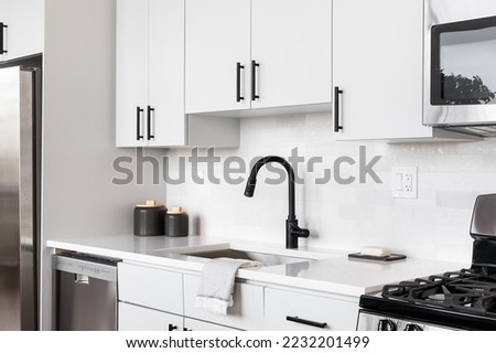 A beautiful white kitchen detail shot with a tiled backsplash, white cabinets, stainless steel appliances, and black hardware and faucet. Royalty-Free Stock Photo #2232201499