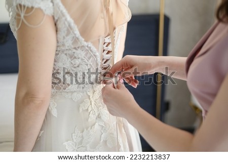 The bridesmaid helps the bride button up her white wedding dress. The bride puts on her wedding dress and prepares to meet the groom. Morning of the bride.Wedding day.