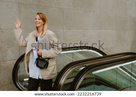 Young beautiful smiling lady in blazer with laptop looking aside and waving , while getting off the escalator